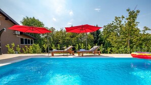 Una piscina con dos sillas de descanso y sombrillas rojas, rodeada de vegetación.