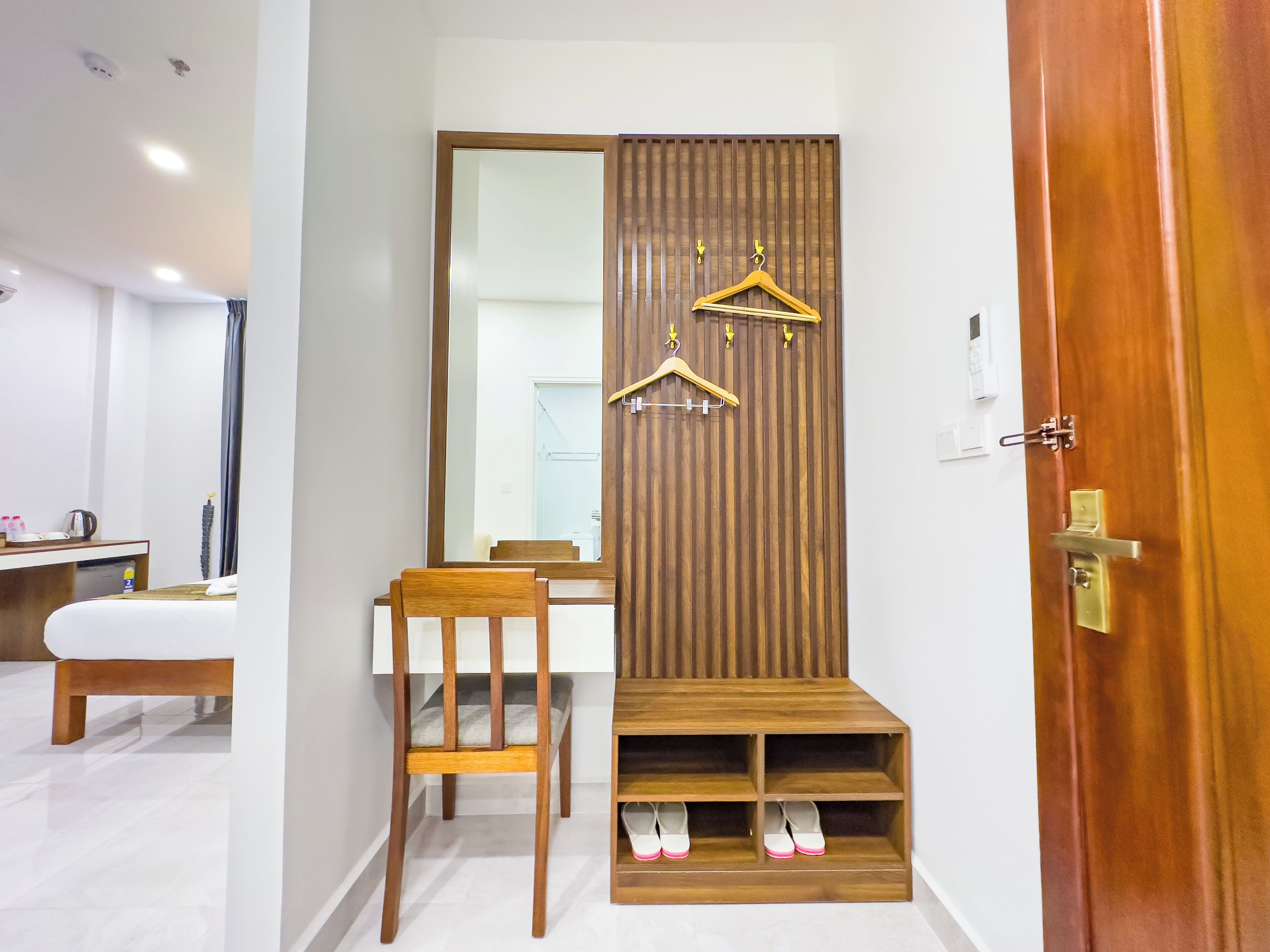 A hotel room with a wooden vanity, a mirror, a wooden shoe rack, a wooden chair, and a white bedside table.