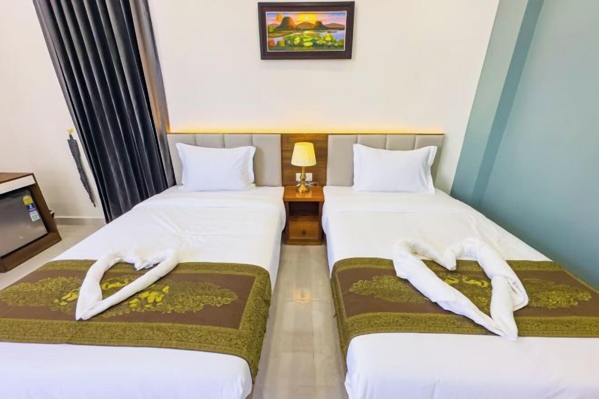 A hotel room with two beds, a nightstand with a lamp, a painting on the wall, and a window with curtains.