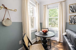 A room with a round table, two chairs, a sofa, a window with curtains, a wall-mounted basket, and a wall art piece.