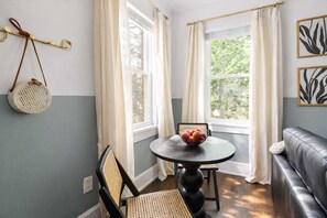 A room with a round table, two chairs, a sofa, a window with curtains, a wall-mounted basket, and a wall art piece.