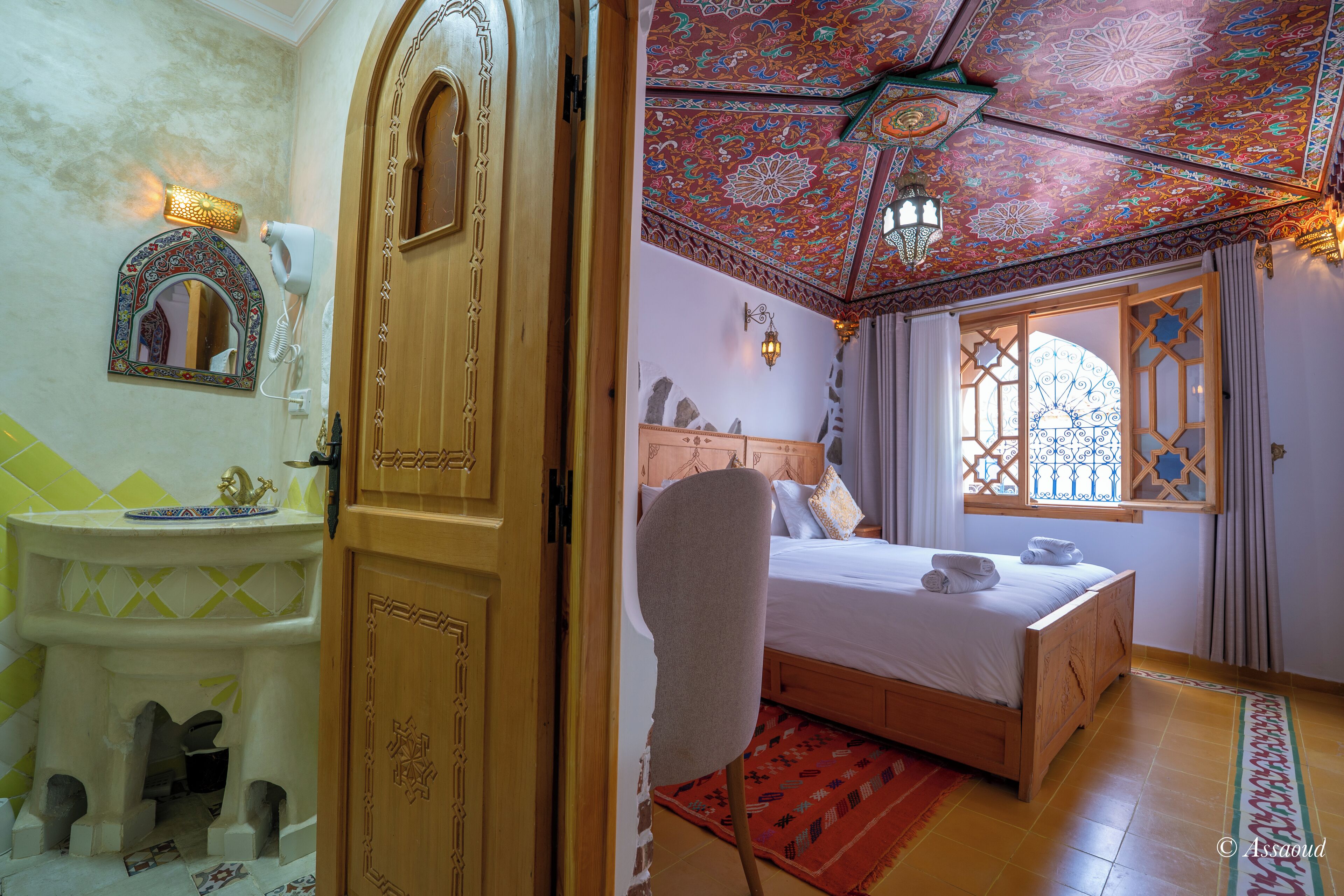 A bedroom with a wooden door, a bed with white linens, a chair, and a decorative ceiling.
