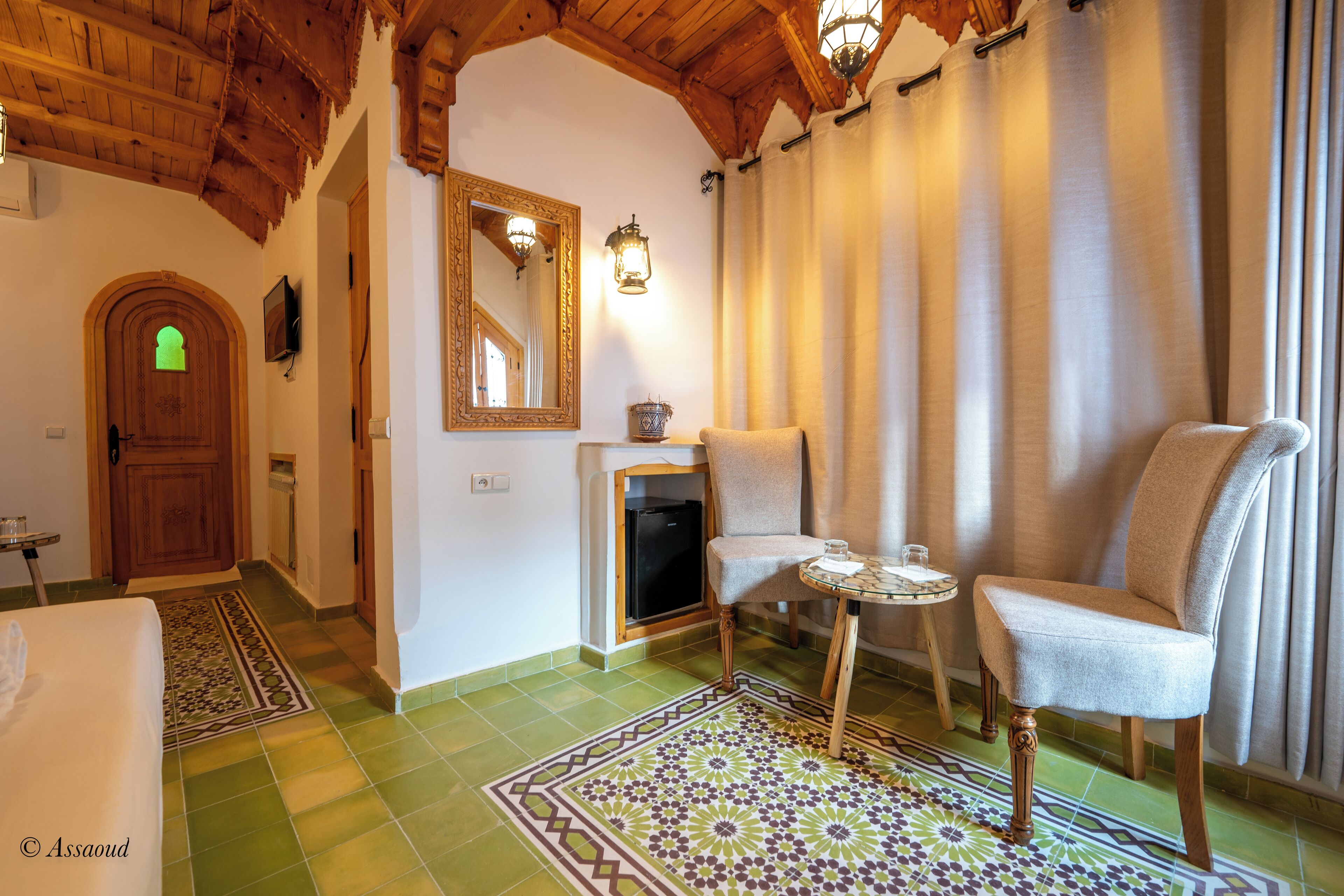 A cozy room with a wooden ceiling, a patterned rug, a chair, a small table, and a door with a green window.