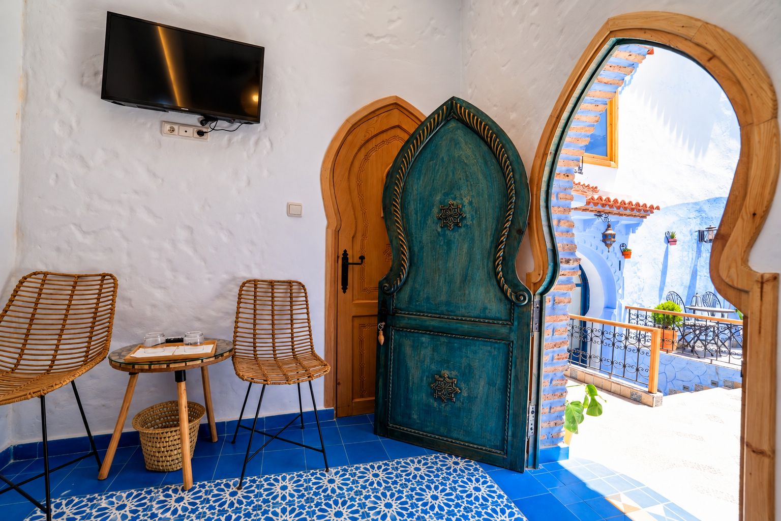Una habitación con un televisor montado en la pared, dos sillas de mimbre, una mesita y una puerta decorativa.