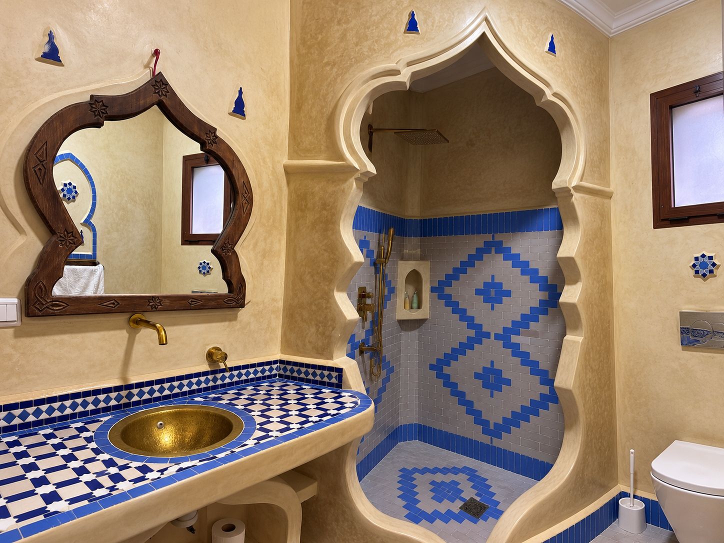 Ein Badezimmer mit einem Becken aus Schachbrettfliesen, einem Spiegel mit Holzrahmen und einer Duschkabine mit blau-weißen Fliesen.