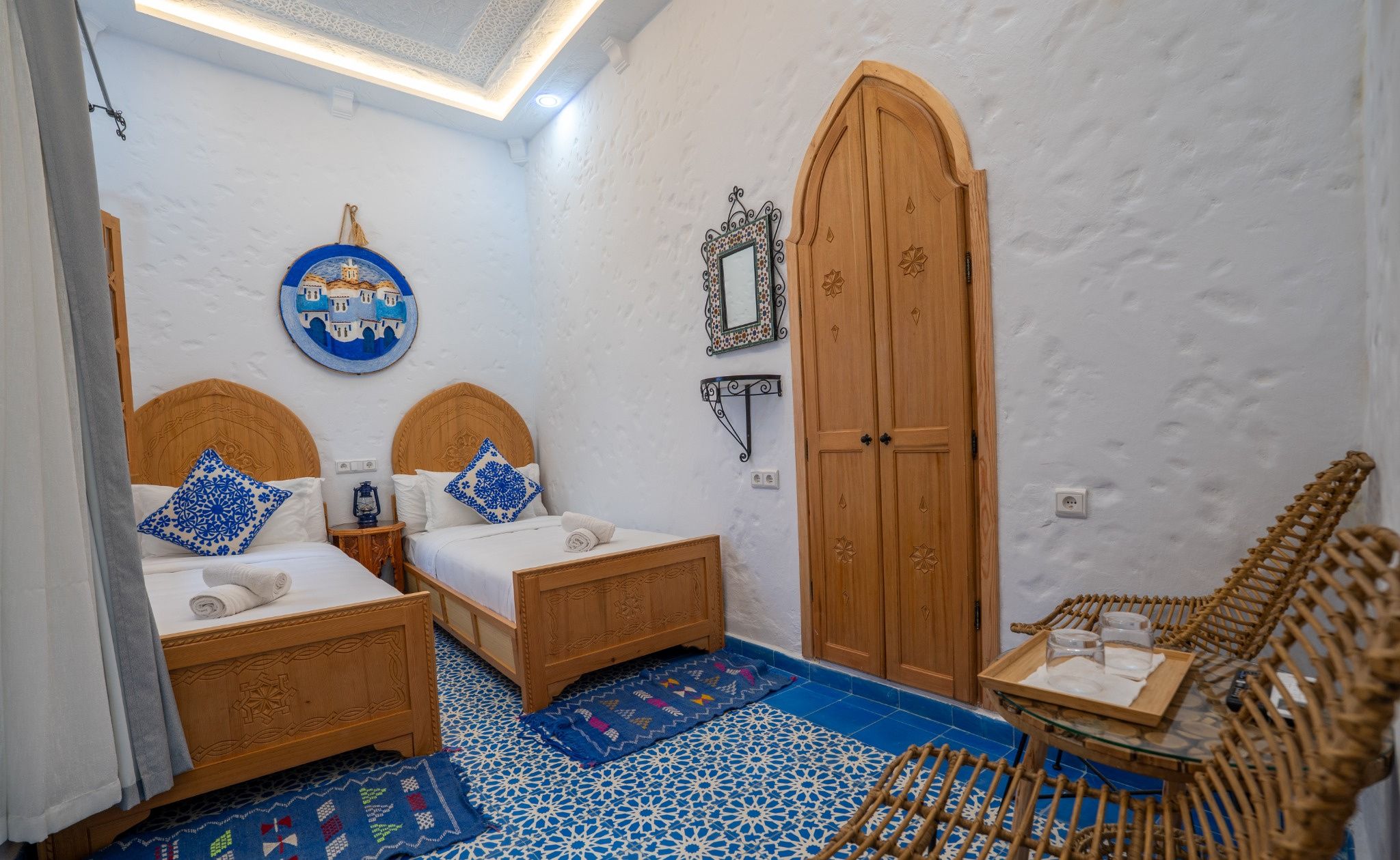 Una habitación con dos camas de madera, un amplio zaguán de madera, una mesita con una silla de mimbre y un suelo con diseño.