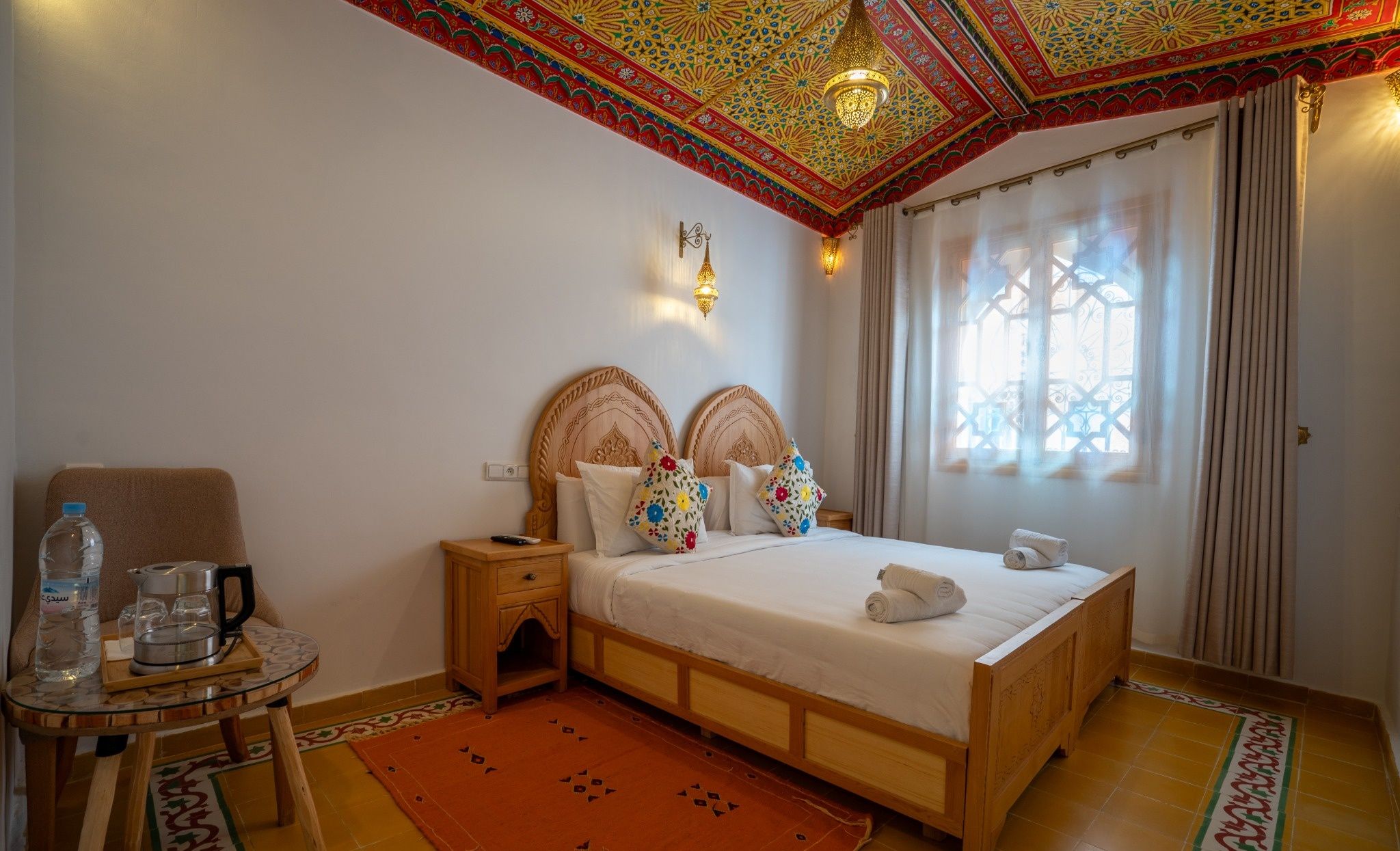 A hotel room with a wooden bed, a small bedside table, a chair, a water bottle, a kettle, and a patterned rug.