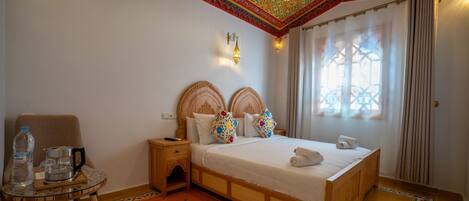 A hotel room with a wooden bed, a small bedside table, a chair, a water bottle, a kettle, and a patterned rug.