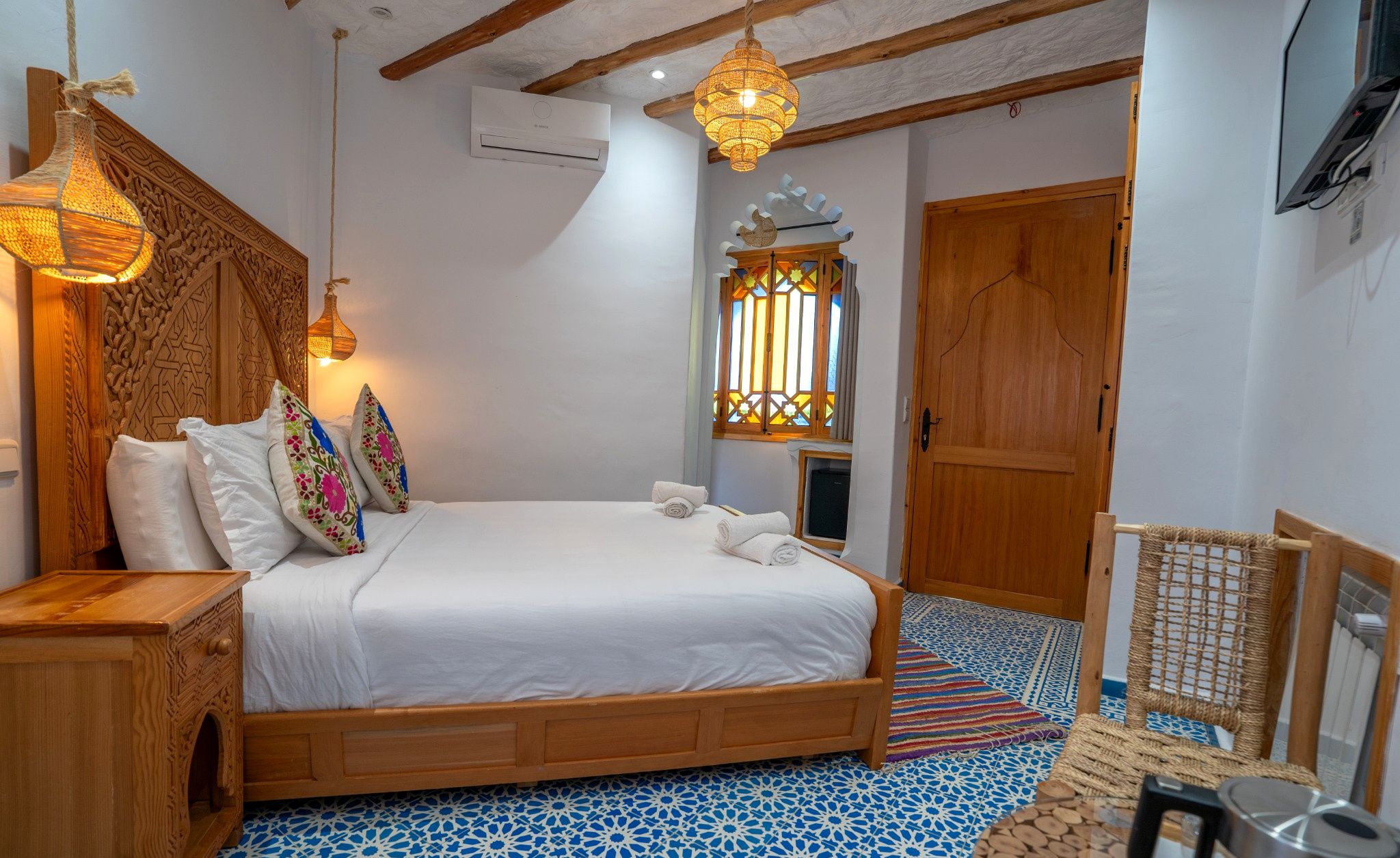 Un dormitorio con cama de madera, almohadones decorativos, una lámpara colgante, una ventana con reja ornamental y una puerta.