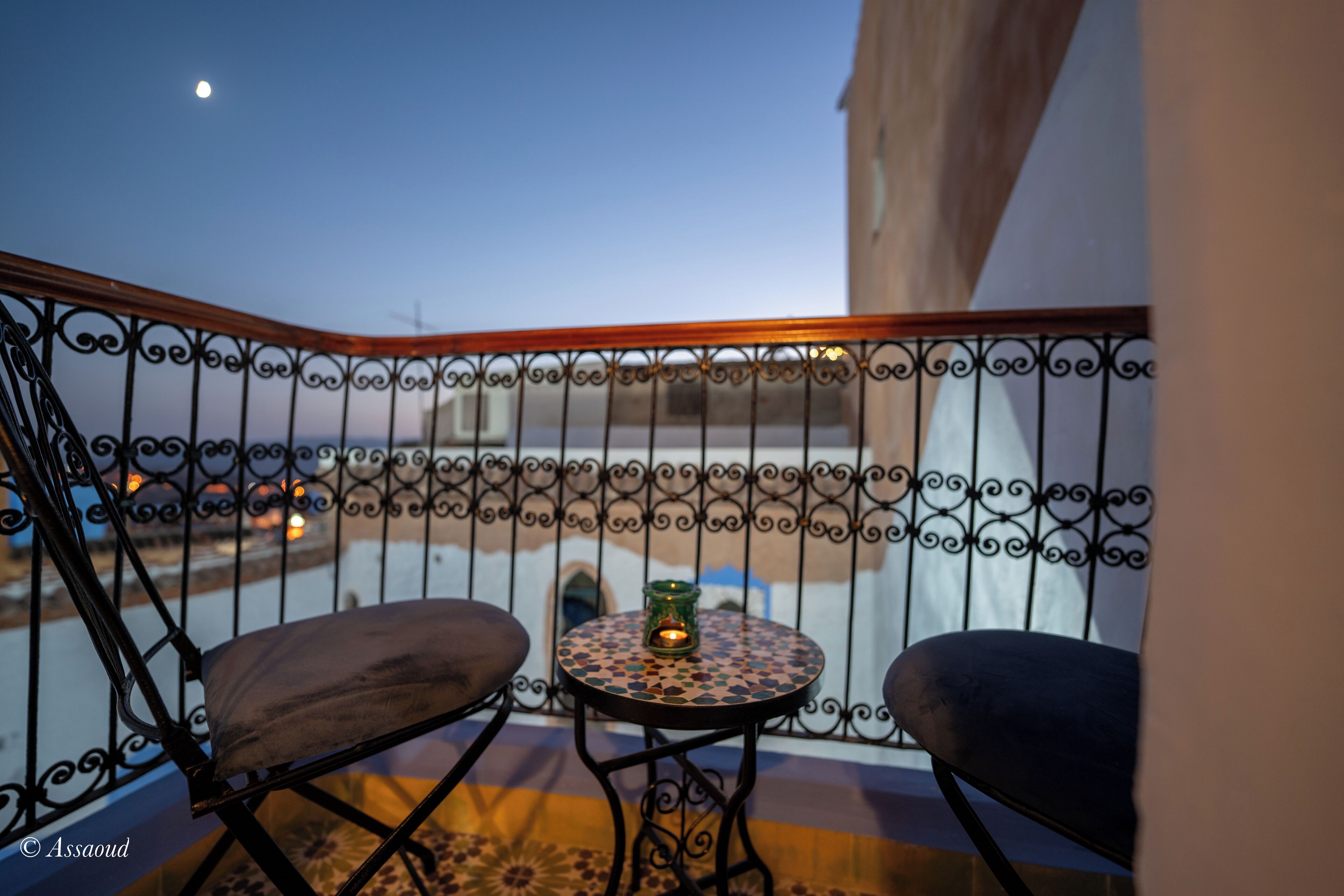 Un balcone con ringhiera in ferro battuto, due poltrone imbottite, un tavolino rotondo con una candela e vista sulla città al crepuscolo.
