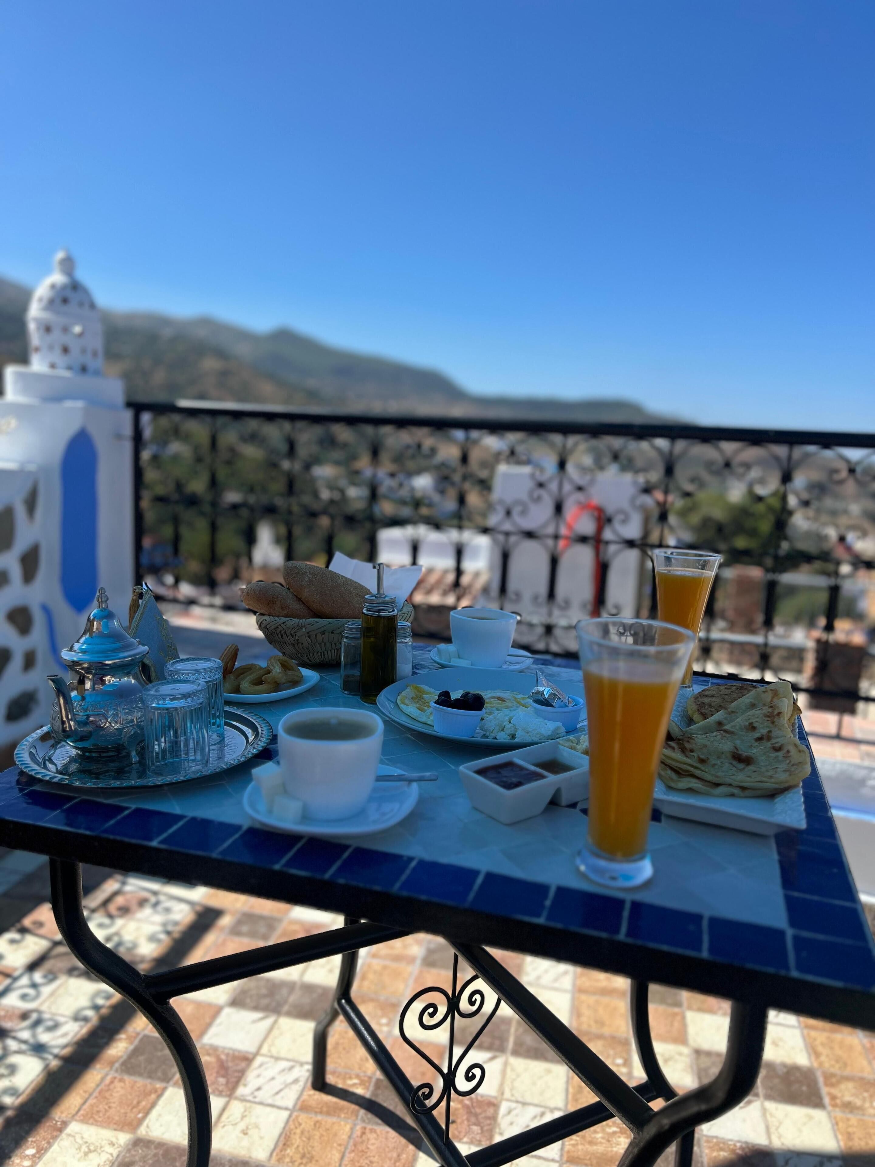 Una mesa con elementos de desayuno, como pan, pasteles y un vaso de zumo de naranja, en un balcón con vistas a la ciudad y las montañas.