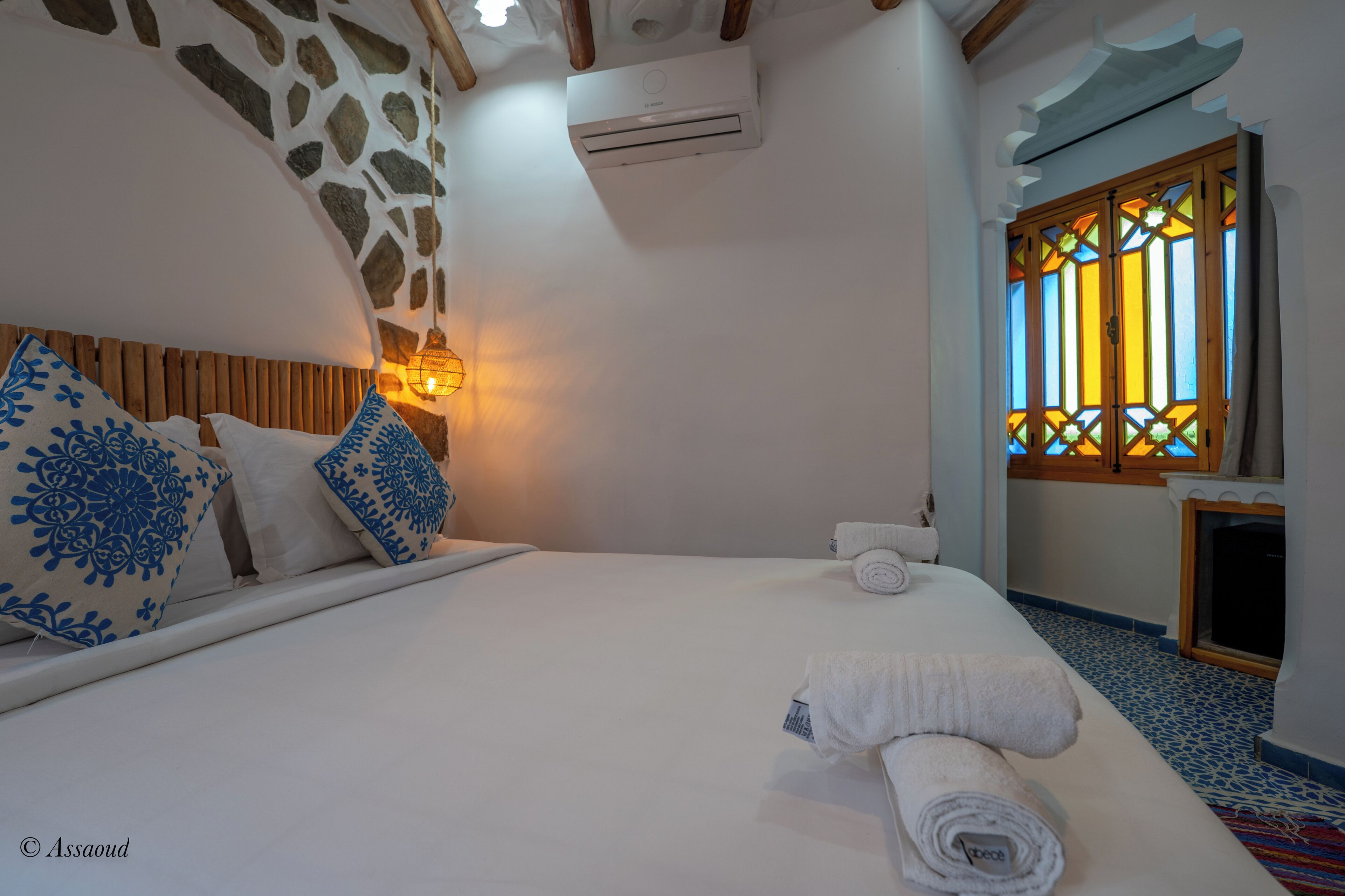 A neatly made bed with white linens and rolled towels, a stone wall with a wooden beam, and a window with stained glass.