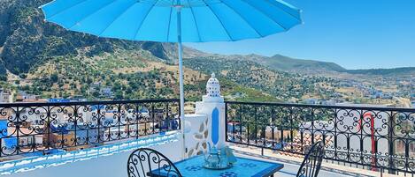 Una terraza con mesa y sillas, un paraguas azul y vistas a un paisaje montañoso.