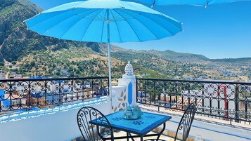 Una terraza con mesa y sillas, un paraguas azul y vistas a un paisaje montañoso.