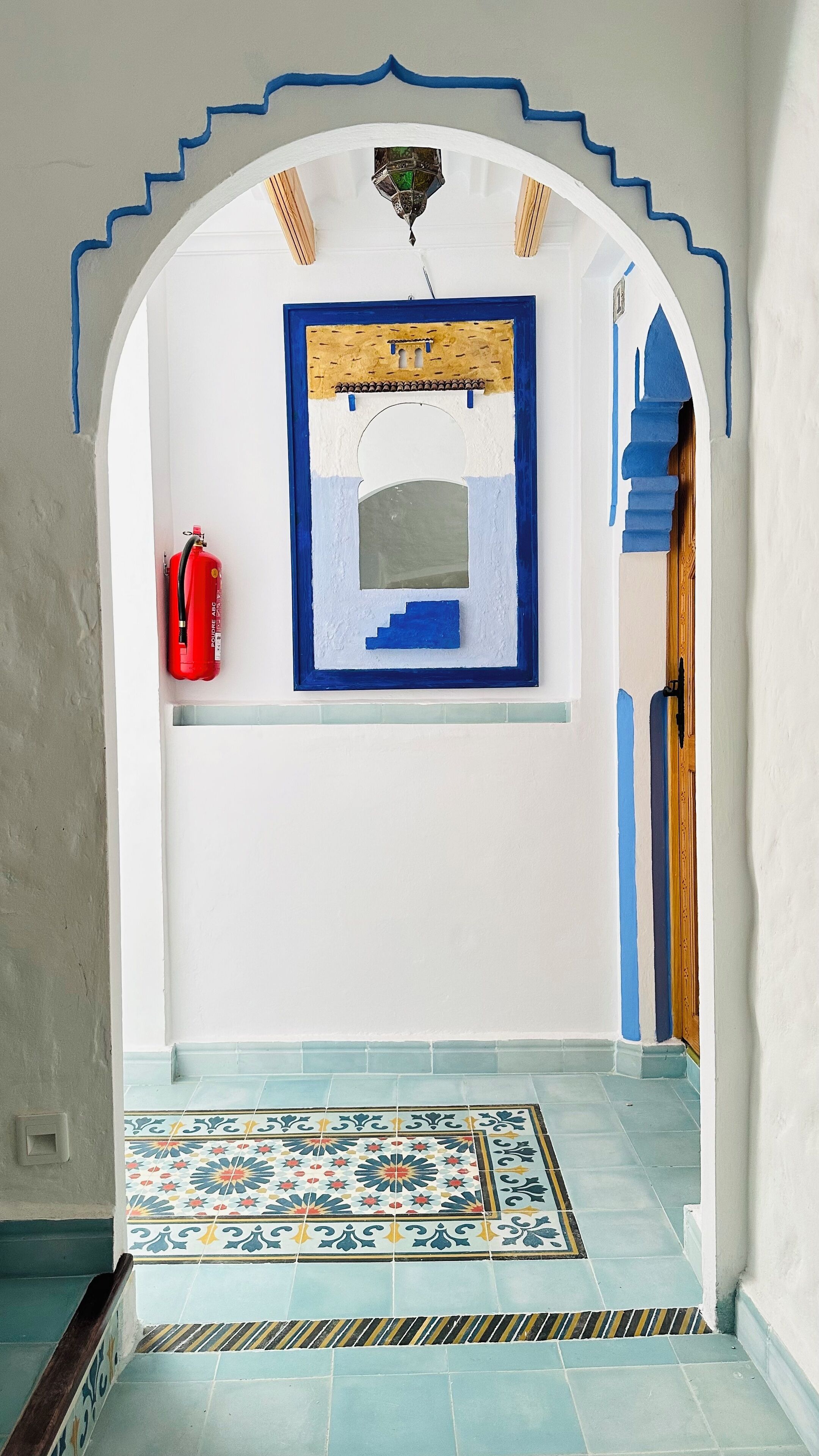 Un pasillo interior con un arco decorativo, un extintor, un suelo de baldosas estampadas y una puerta de paneles de madera.