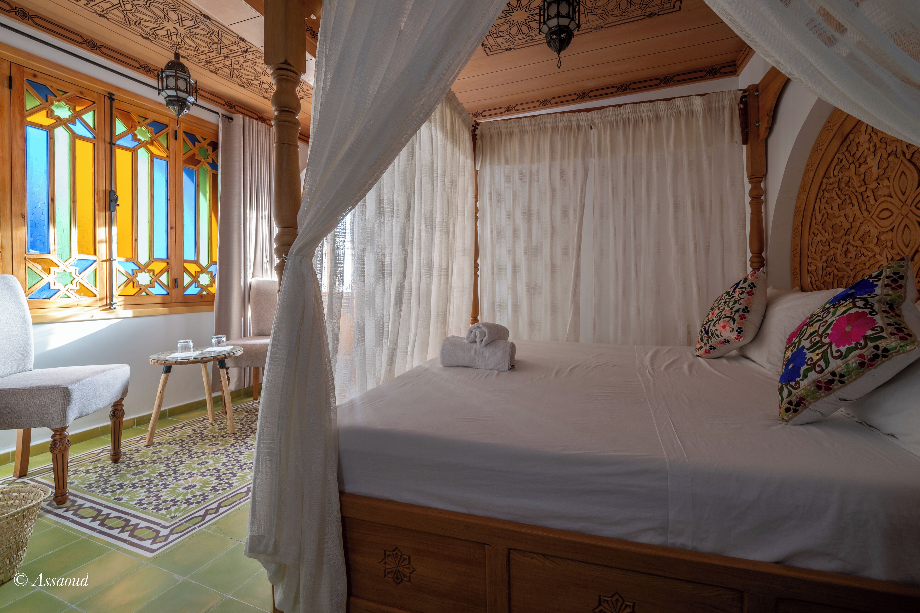A bedroom with a four-poster bed, a chair, a small table, and a decorative rug.