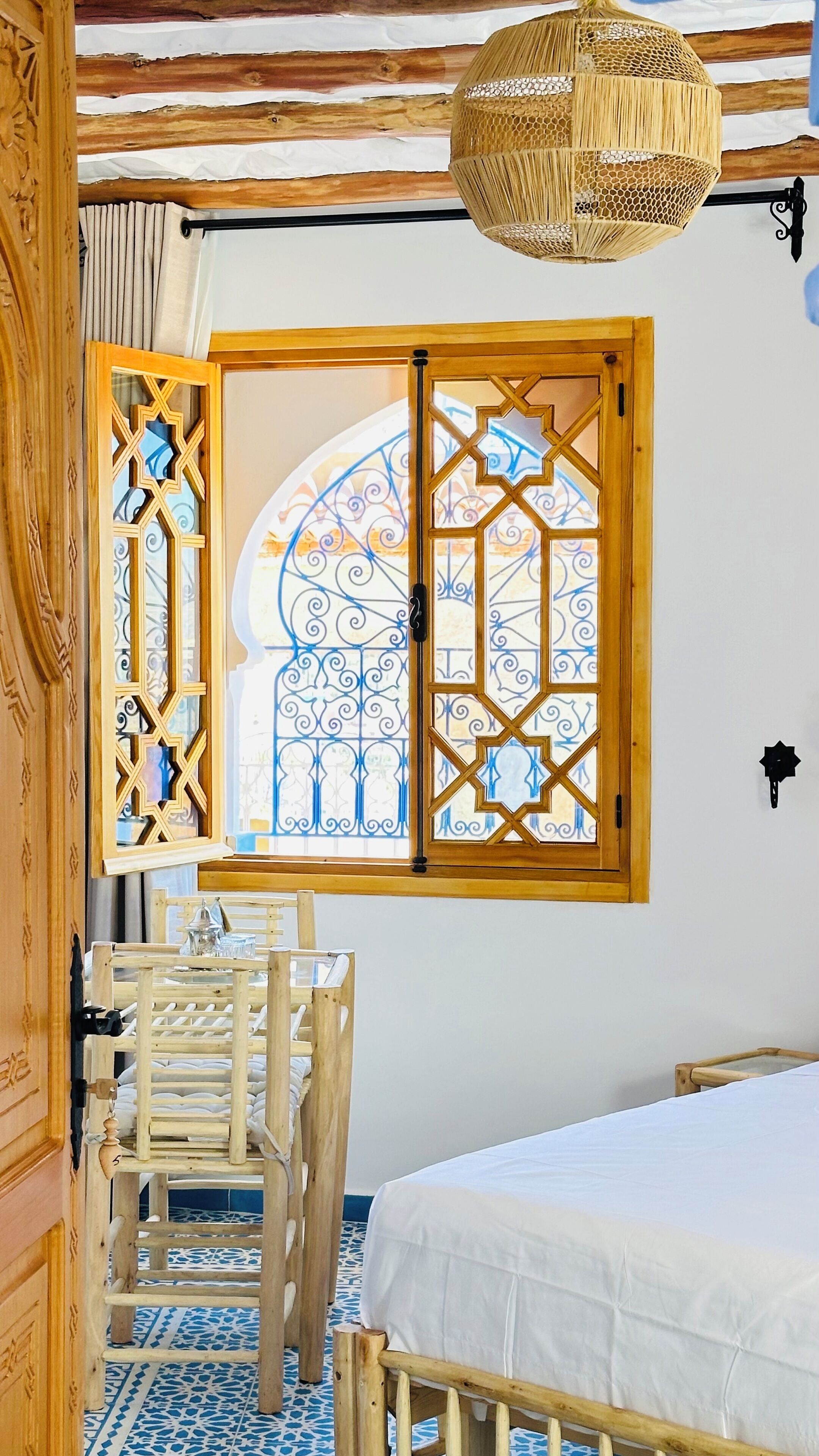 Una habitación con una cama, una silla de madera, una lámpara colgante de ratán y una ventana con ornamentación metálica detallada.