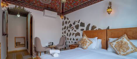 A hotel room with a large bed, a sitting area with a chair and table, a decorative ceiling, and a stone wall.