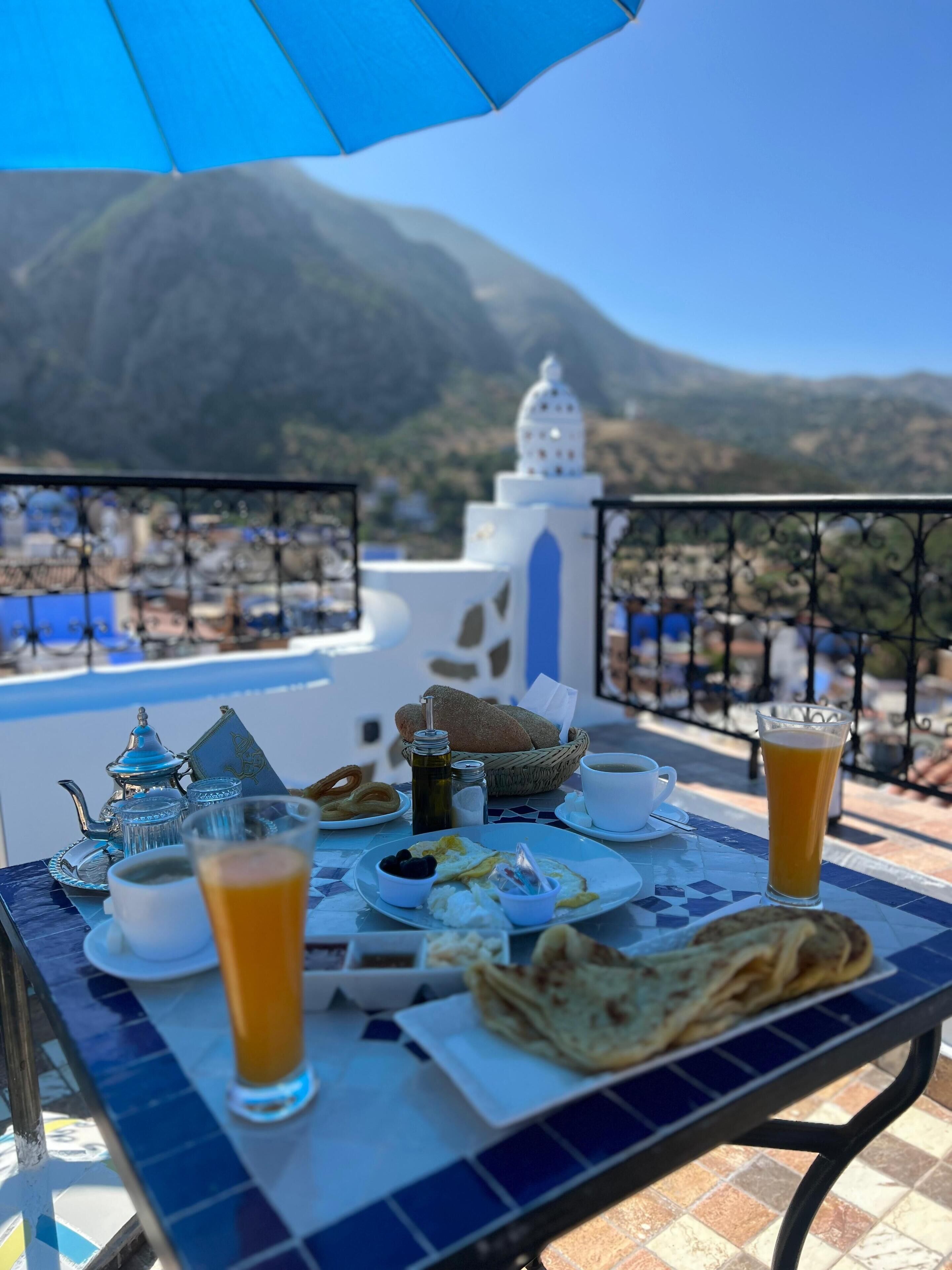 Una mesa con una preparación de comida que incluye pan, un hervidor de té, tazas y un vaso de zumo de naranja, con un paisaje montañoso de fondo.