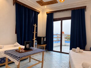 A hotel room with two beds, a table with a striped tablecloth, a kettle, a bottle of water, a TV, and a view of the sea through a large window. - Alhambra Ryad & Spa (Chefchaouen)