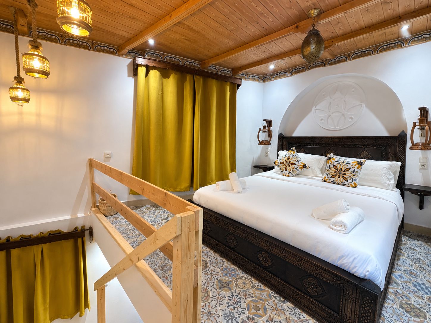 Ein Hotelzimmer mit einem Bettgestell aus Holz, einem großen Bett mit weißer Bettwäsche, dekorativen Kissen und einem Holz-Kopfteil sowie einem Fußboden mit Muster.