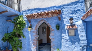 Un callejón estrecho con un edificio azul, una puerta de madera y un pequeño patio con una silla y plantas en macetas.