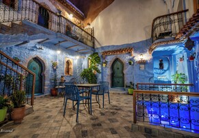 A courtyard with a tiled floor, blue-painted walls, a wooden staircase, and a dining area with blue chairs and a table.