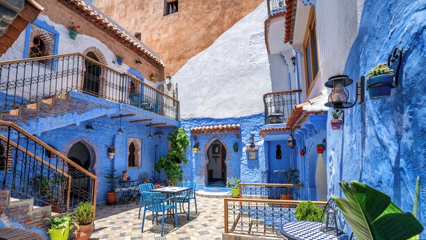 Un patio con edificios de color azul, mobiliario exterior y plantas en macetas.