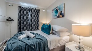 A neatly made bed with a blue blanket, white pillows, and a teal throw pillow. There is a white bedside table with a lamp and a painting on the wall.