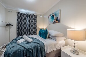 A neatly made bed with a blue blanket, white pillows, and a teal throw pillow. There is a white bedside table with a lamp and a painting on the wall.
