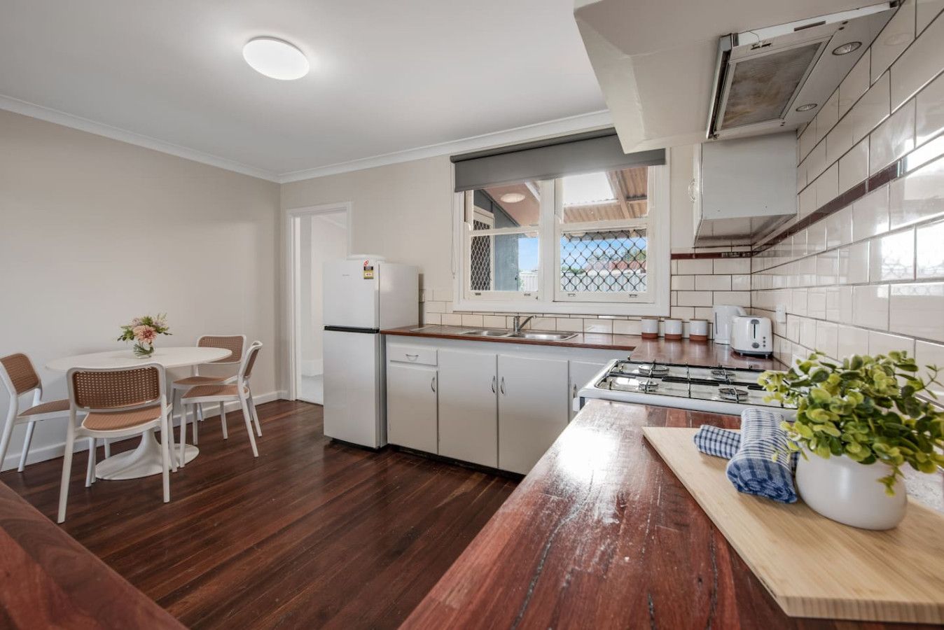 A modern kitchen with a dining area, wooden flooring, white cabinets, and a window with a view.