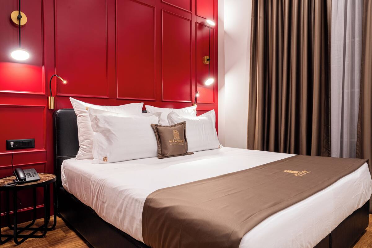 Une chambre d’hôtel avec un mur rouge, un lit avec une literie blanche et marron, une table de chevet avec un téléphone et une fenêtre avec des rideaux.