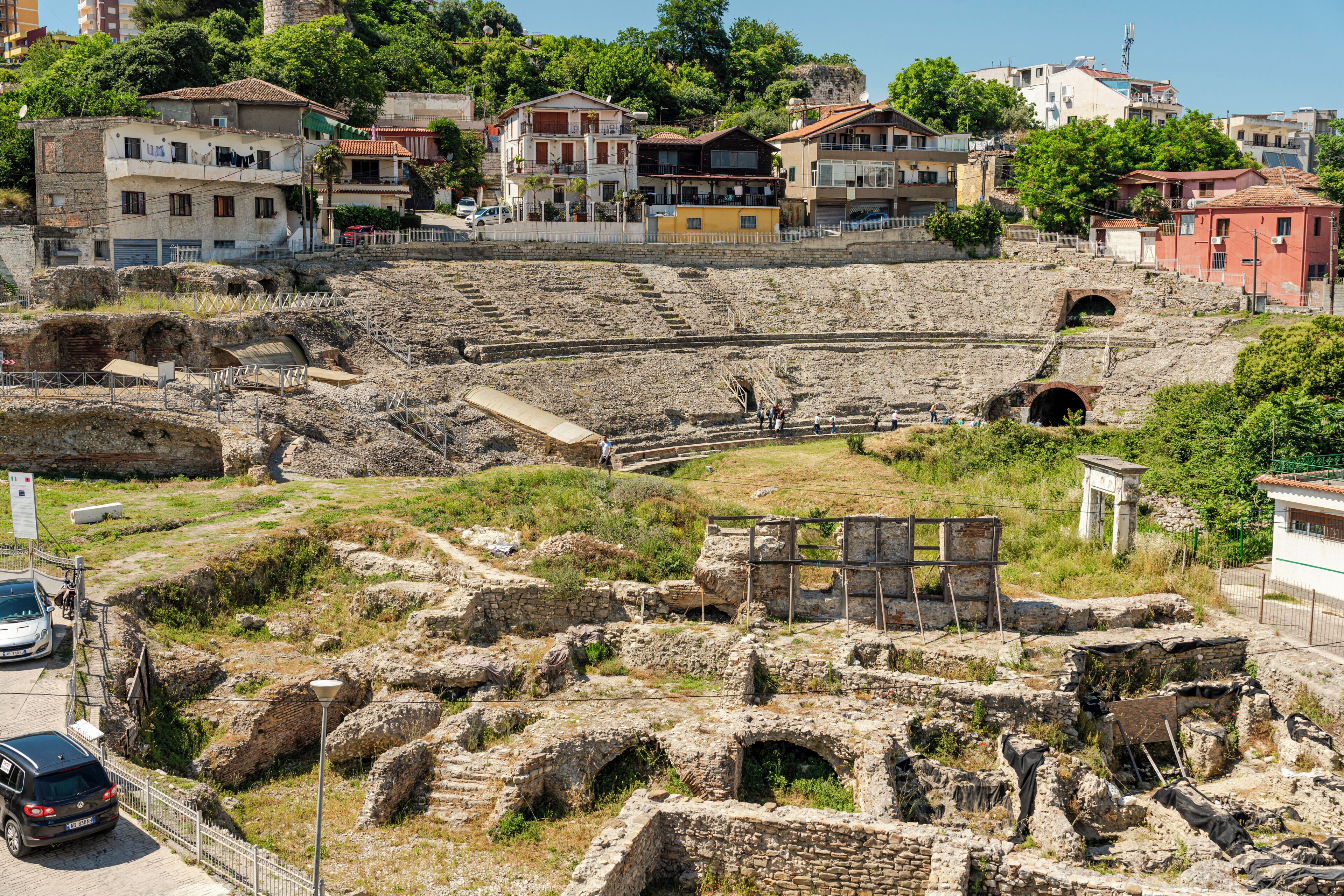 Alte Ruinen mit Steinbauten, einem teilweise erhaltenen Amphitheater und modernen Gebäuden im Hintergrund.