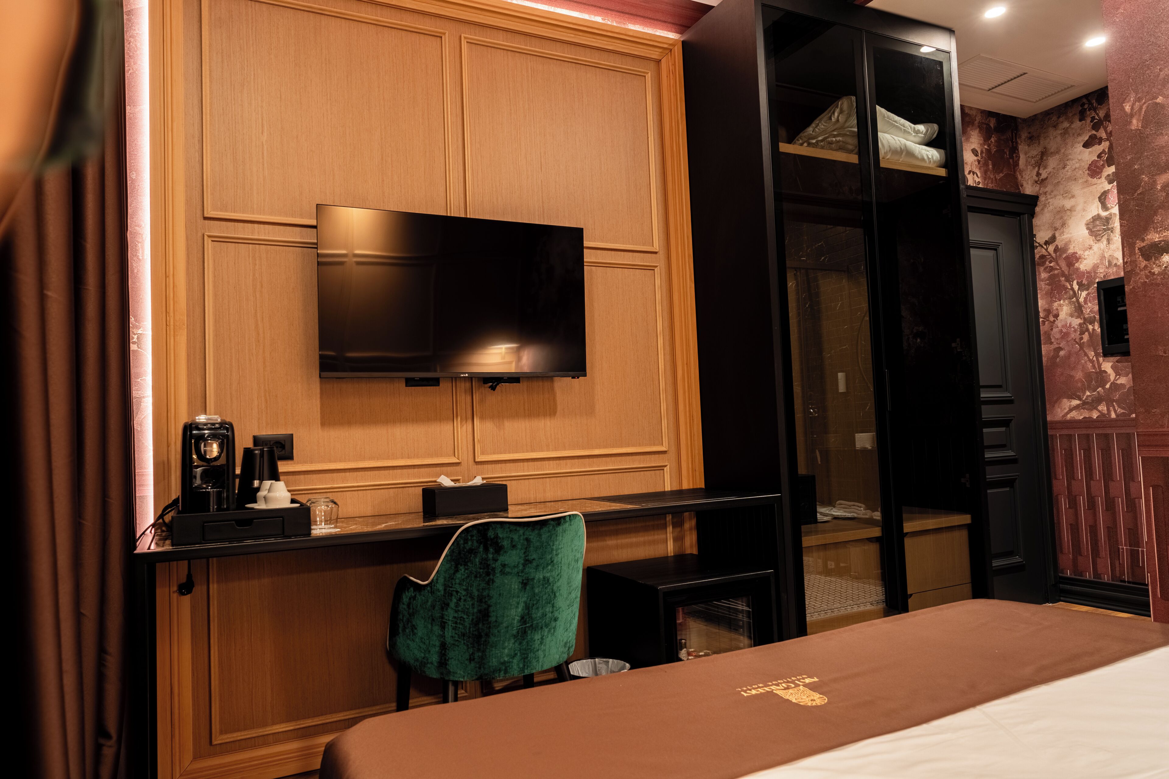 A hotel room with a bed, a TV mounted on a wooden panel wall, a desk with a chair, and a wardrobe.