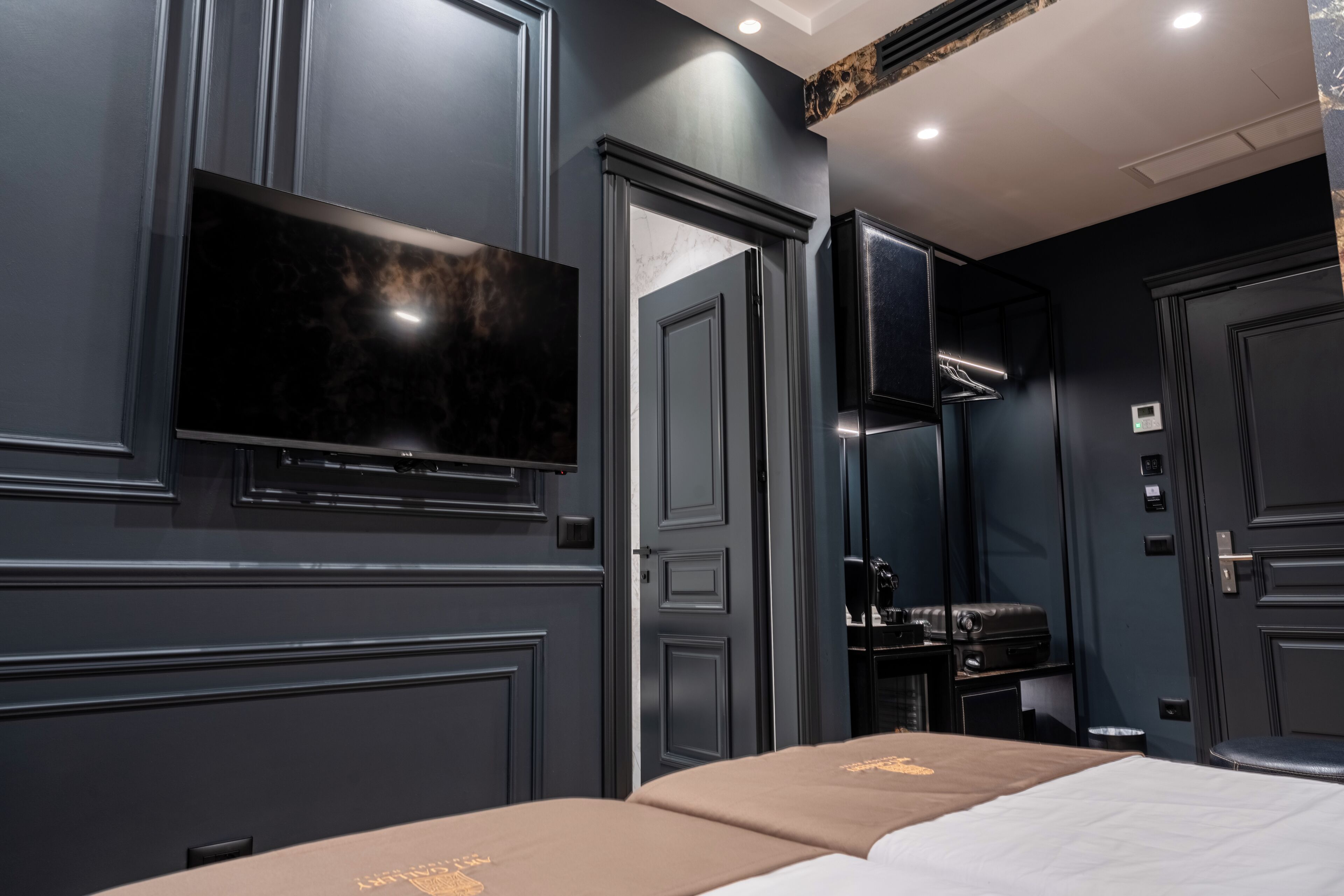 Une chambre d’hôtel moderne dotée d’un grand téléviseur à écran plat fixé au mur, d’un lit recouvert d’une couvre-lit beige et d’une armoire de couleur sombre.