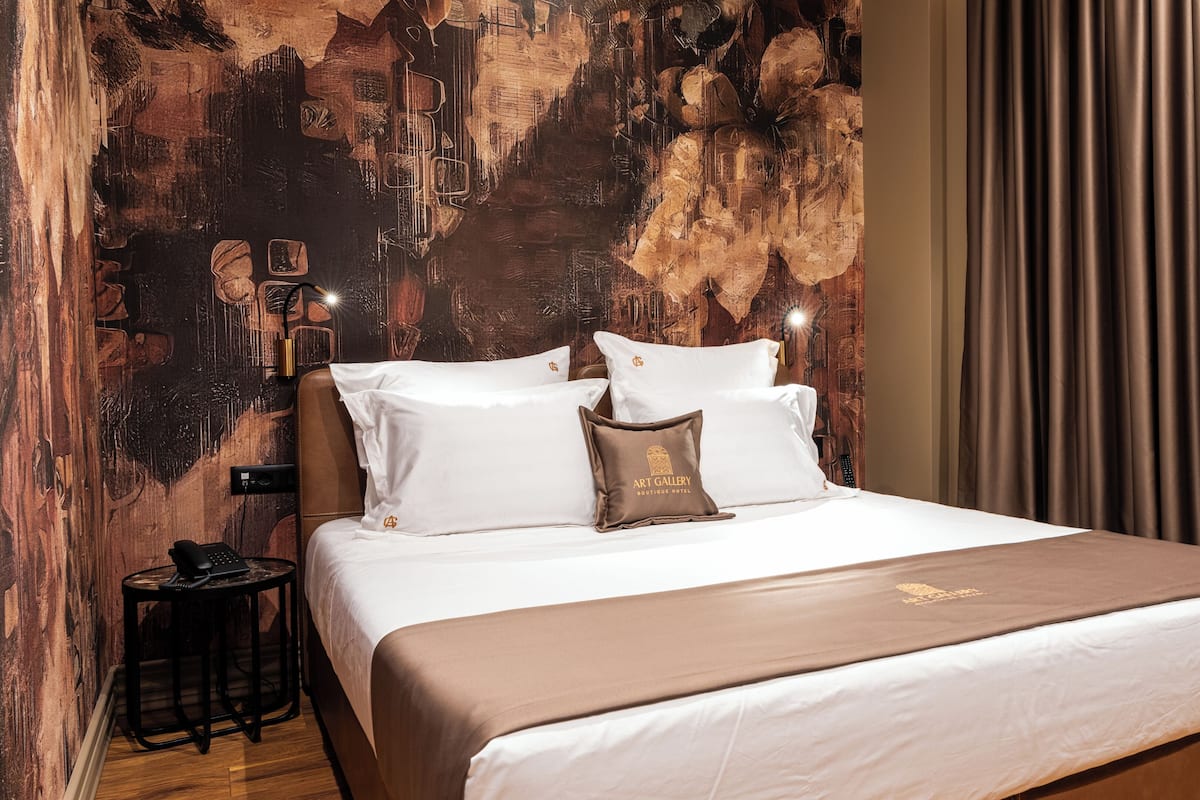 Une chambre d’hôtel avec un grand lit, une table de chevet, une lampe et un mur orné d’une fresque à motifs.