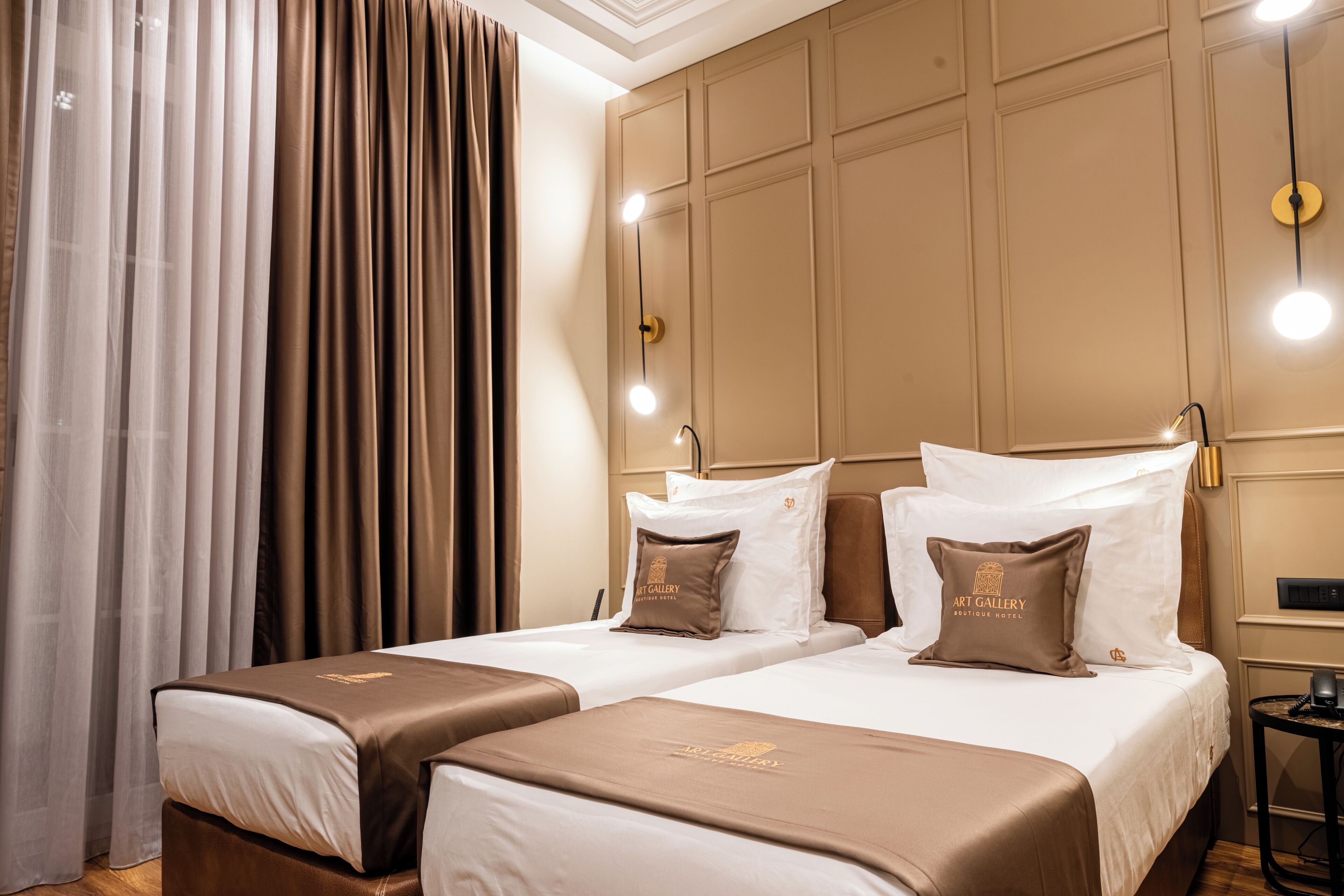 Une chambre d’hôtel avec deux lits, une grande fenêtre avec des rideaux, un luminaire fixé au mur et une petite table avec un téléphone.