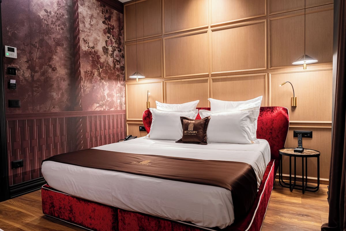 Une chambre d’hôtel avec un grand lit, une tête de lit rouge rembourrée, un panneau de commande fixé au mur et une petite table ronde avec une lampe.