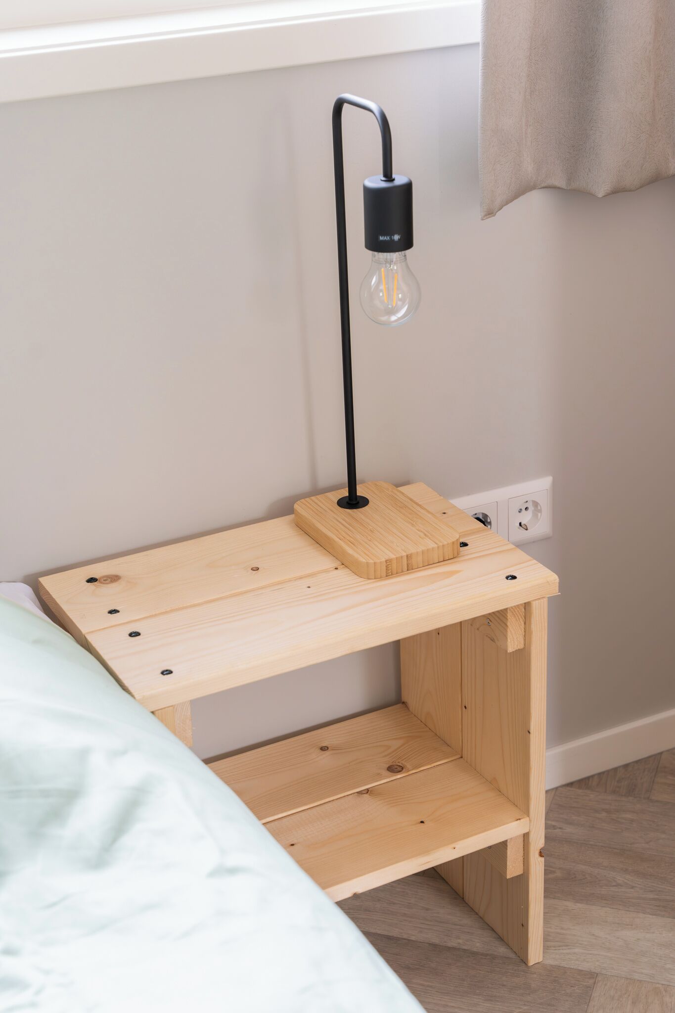 A wooden bedside table with a black lamp and a light bulb, positioned next to a bed with a light green bedspread.