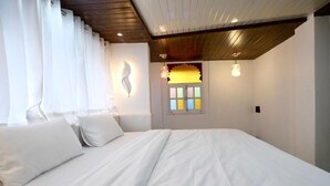 Un dormitori amb un llit gran, roba de llit blanca, una finestra amb vitralls de colors i un sostre de fusta.