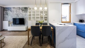 A modern living room with a TV, dining table, and chairs, a kitchen with blue cabinets, and a large window. - Native Apartments Orawska 14 (Kraków)