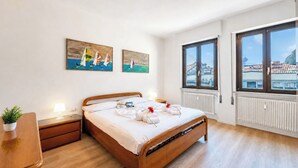 A hotel room with a wooden bed, a nightstand, a potted plant, and two paintings on the wall. - Casa Lucia in Old Riva Town With Balcony View (Riva del Garda)