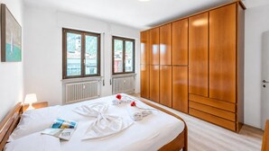 Une chambre d’hôtel avec un lit, une armoire, deux fenêtres, un radiateur et une lampe.