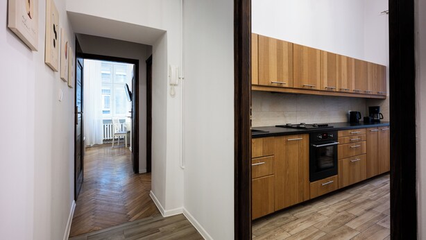A modern kitchen with wooden cabinets, a black countertop, and an oven. A hallway with wooden flooring leads to a dining area with a white chair and table. - Native Apartments Skawińska 5 (Kraków)