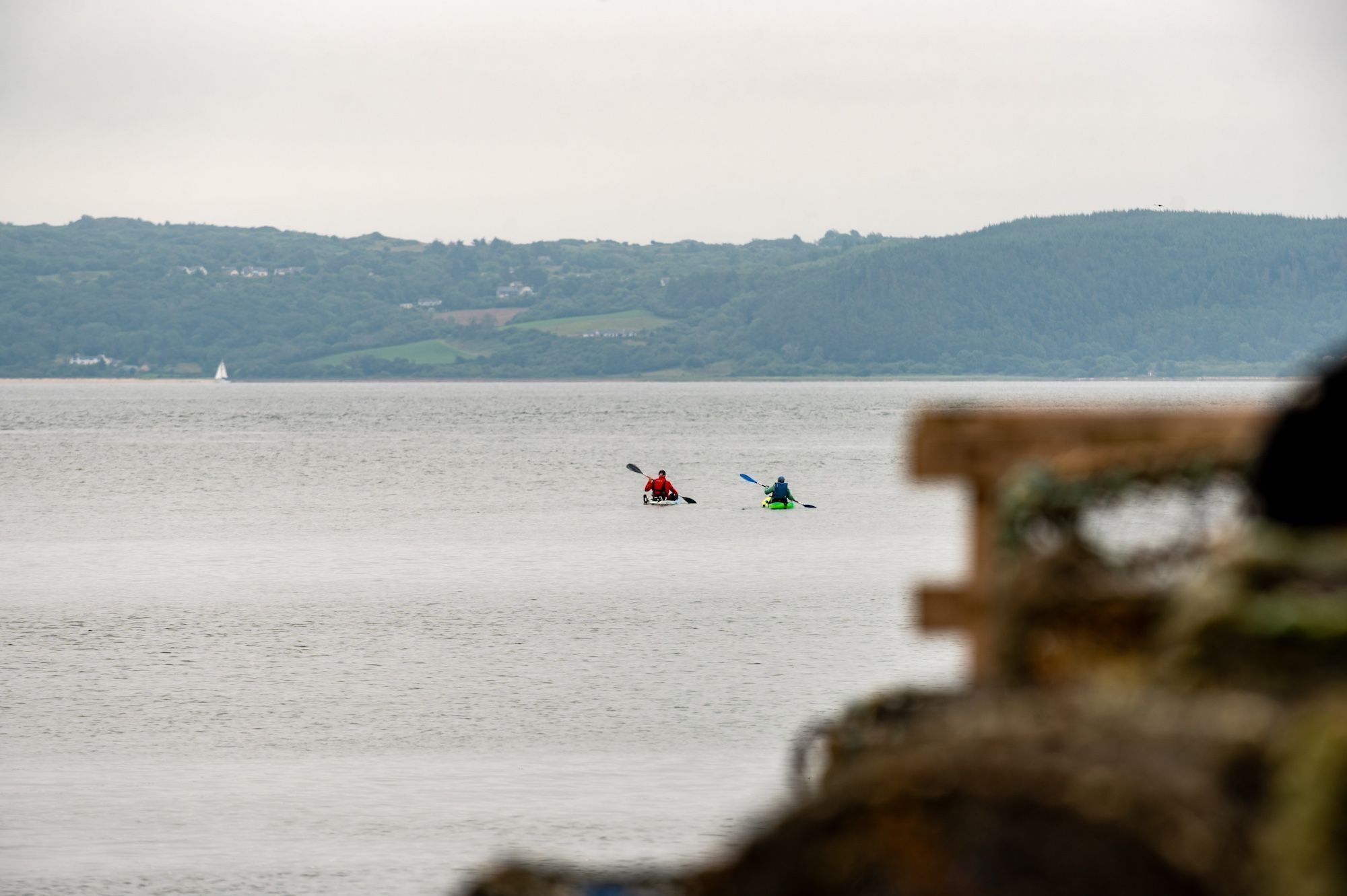 A calm lake with kayakers in the distance and a sailboat on the horizon.