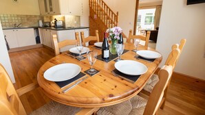 Un coin repas avec une table en bois dressée pour quatre personnes, entourée de chaises en bois. On aperçoit un escalier au fond, menant à un autre niveau.