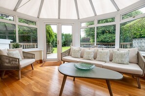 A conservatory with a glass door leading to a garden, a sofa set with patterned cushions, and a glass bowl on a table.