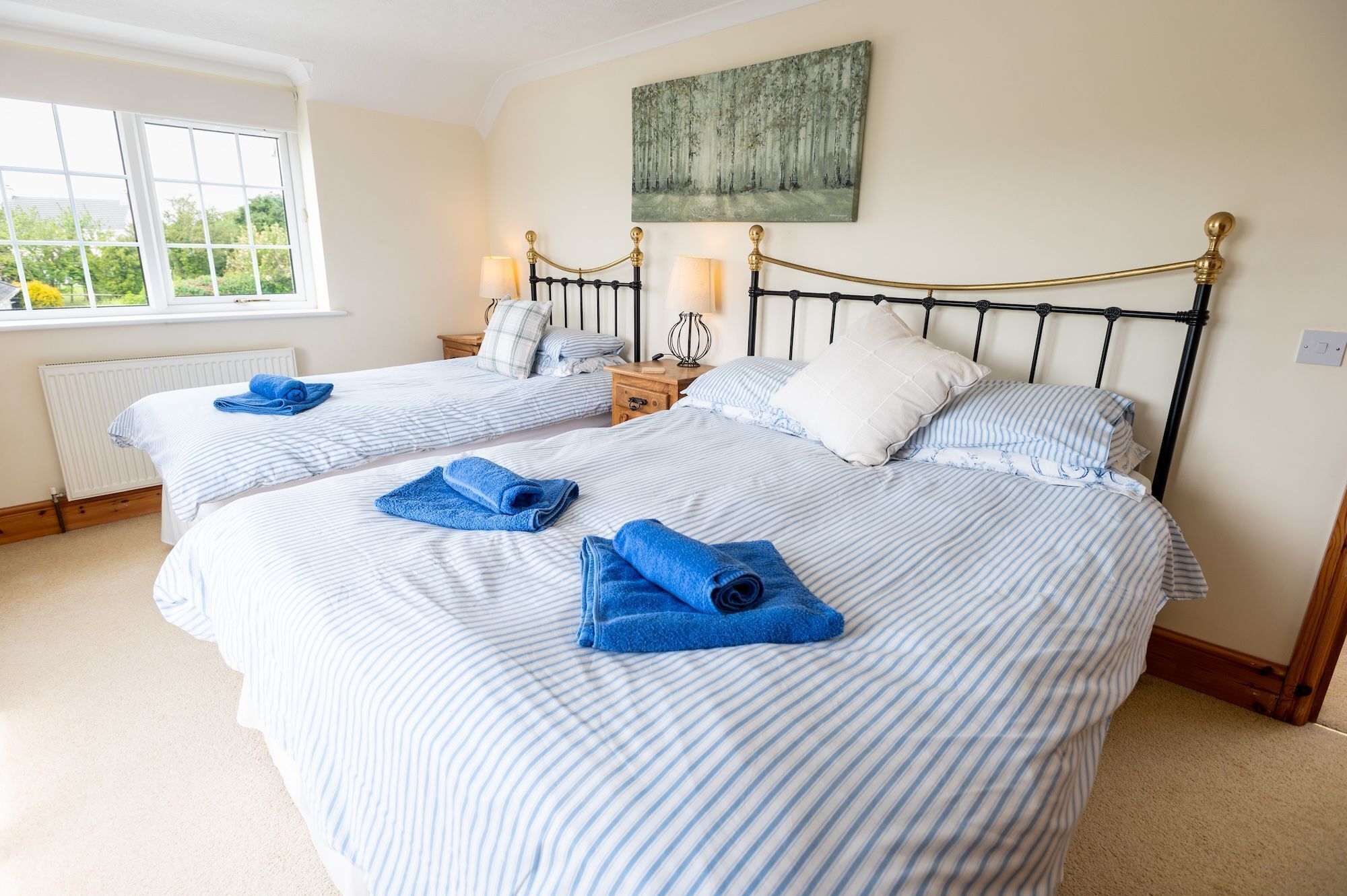 Habitación de hotel con dos camas, cada una con ropa de cama a rayas azules y toallas enrolladas. Cuenta con una ventana que proporciona luz natural, un radiador y un cuadro en la pared.