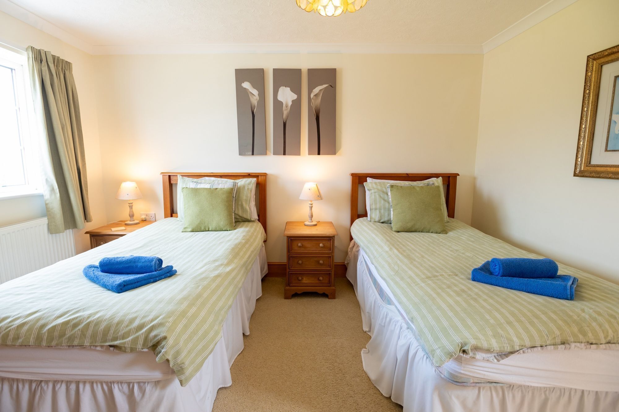 Habitación doble con dos camas individuales, cada una con una colcha verde a rayas, una almohada blanca y una toalla azul. Hay mesitas de noche con lámparas, una ventana con cortinas y un cuadro enmarcado en la pared.