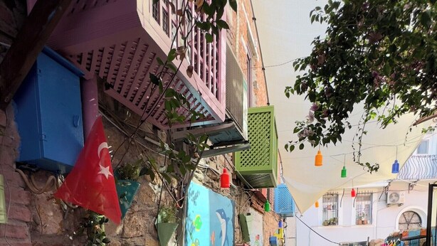 Une rue pavée, avec des tables et des chaises colorées disposées à l’extérieur, et un bâtiment doté d’un balcon.